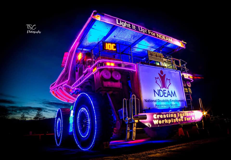 A photograph of a giant iron ore haul truck lit purple and blue, including the tires and wheel rims. There is a lighted sign across the front of the dumper that says, "Light It Up! For NDEAM". There are also graphics that say National Disability Employment Awareness Month, on the truck.