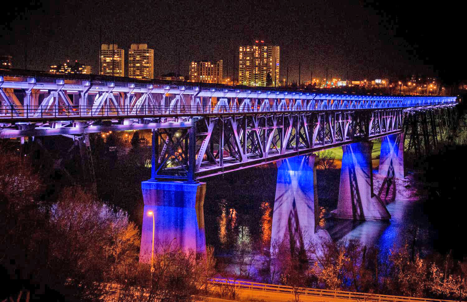 The girders and piers of the High-Level Bridge in Edmonton lit purple and blue, against city skyline.