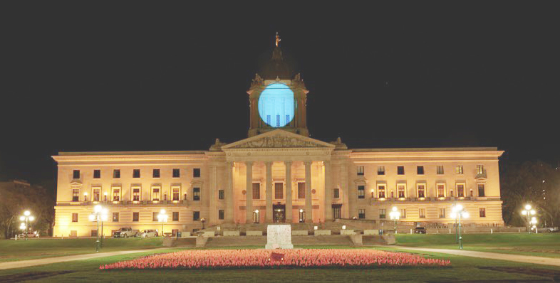 A wide, head-on shot that shows the searchlight-style purple and blue lighting of the dome on the Manitoba Legislature for Light It Up! For NDEAM.