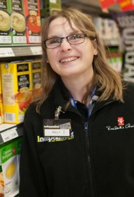 Young white female with shoulder-length brown hair. The person is smiling and looking directly at a camera. The person is wearing glasses and a black sweater with two grocery store logos: on the left, President's Choice is written in white colour; on the right, the word "Independent" is written in white with some yellow. The person is gearing a name tag with the name "Holly". The person is standing next to a grocery aisle displaying shelved items.