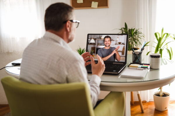A man who works remotely is sitting at his desk having a conversation on a video call with another man in American Sign Language.
